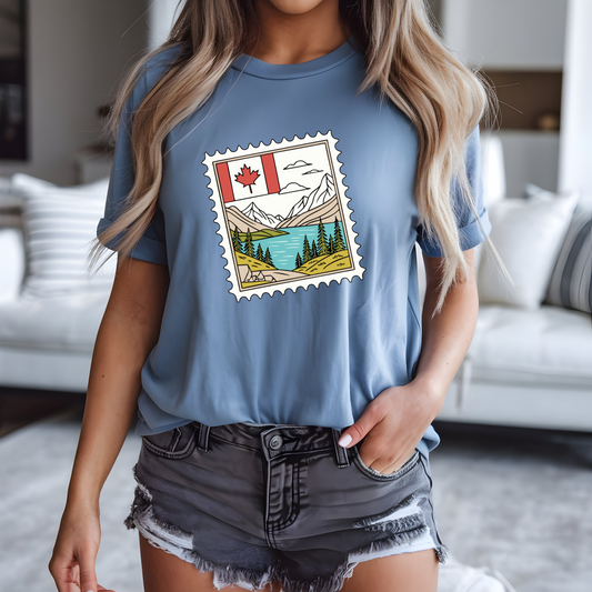 On the front of this custom Canadian pride short sleeve shirt is an image of a Canadian postage stamp with the Canadian flag in one corner and an image of mountains, a lake, and trees the make up the rest of the stamp.