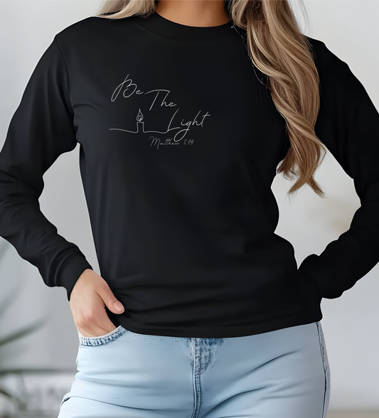 This Christian unisex Long Sleeve t-shirt features the words Be The Light Matthew 5:14 in a simple cursive font with an image of a line drawing of a candle next to the word light.