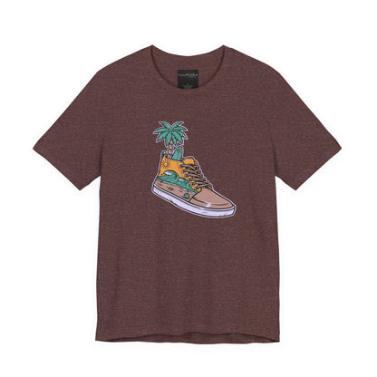 On the front of this custom handmade short sleeve shirt is an image of a high top running shoe with ocean waves running along the side of the shoe, coming out of the top of the shoe where your foot goes is a palm tree and a surf board.