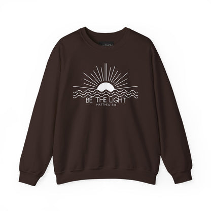 This cozy Christian crewneck sweatshirt features a sunrise over the ocean in the background with the bible verse Be the Light Matthew 5:14 in front of the image.