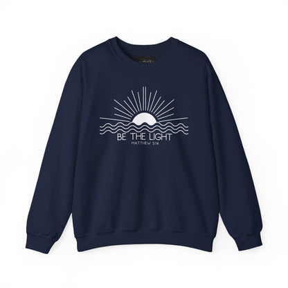 This cozy Christian crewneck sweatshirt features a sunrise over the ocean in the background with the bible verse Be the Light Matthew 5:14 in front of the image.