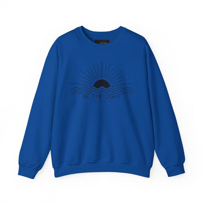 This cozy Christian crewneck sweatshirt features a sunrise over the ocean in the background with the bible verse Be the Light Matthew 5:14 in front of the image.