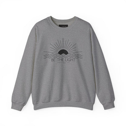 This cozy Christian crewneck sweatshirt features a sunrise over the ocean in the background with the bible verse Be the Light Matthew 5:14 in front of the image.