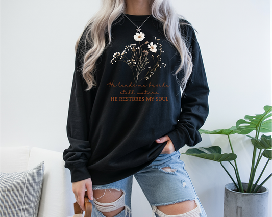 On the front of this women's Christian long sleeve shirt are a bunch of beige and brown flowers and leaves on the top of the shirt and under that are the words He leads me beside still waters in a cursive font with the words He restores my soul underneath that in a modern font.