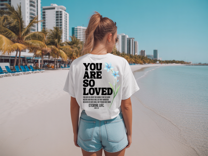 On the back of this Christian custom unisex t-shirt are the words You are loved in large bubble letters with a blue wildflower next to the words, the bible scripture John 3:16 is underneath that reads For God so loved the world that he gave his only son, so that whoever believes in him shall not perish but have everlasting life.