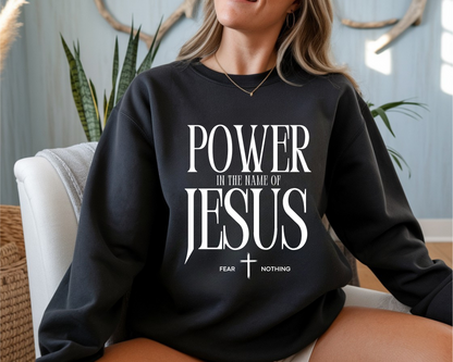 On the front of this Christian pullover sweatshirt it says Power In The Name Of Jesus with a Jesus cross underneath it, on either side of the cross are the words Fear and Nothing.