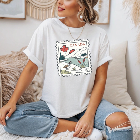 On the front of this custom short sleeve shirt is an image of a Canadian postage stamp, there is a maple leaf, the rocky mountains, a lake and a Canadian goose on the postage stamp.
