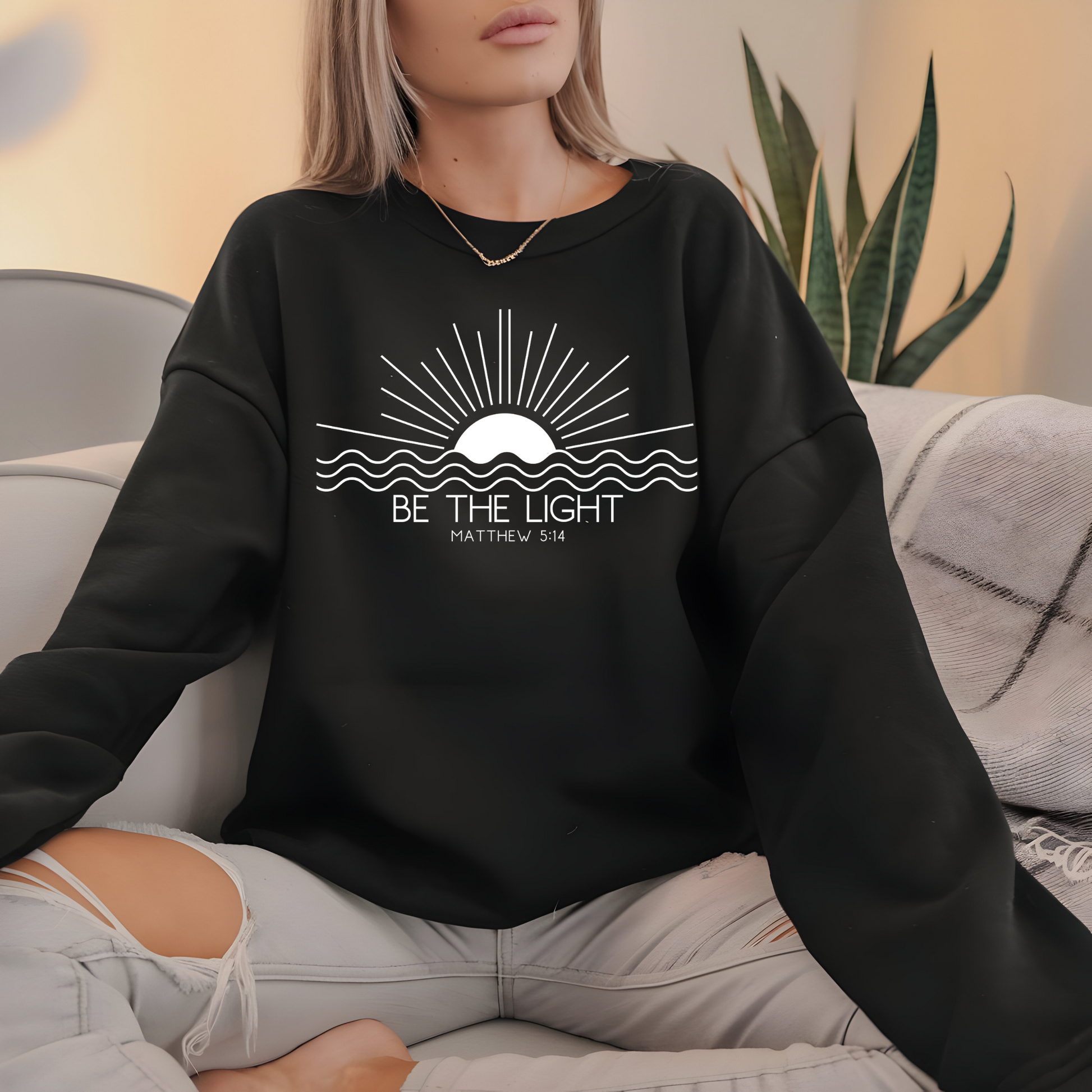 This cozy Christian crewneck sweatshirt features a sunrise over the ocean in the background with the bible verse Be the Light Matthew 5:14 in front of the image.