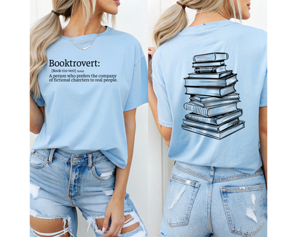 On the front of this custom unisex short sleeve shirt is the word booktrovert with the definition of the word underneath, on the back of the shirt is an image of a large stack of books.
