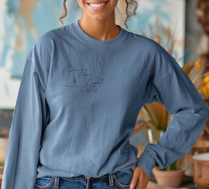 This Christian unisex Long Sleeve t-shirt features the words Be The Light Matthew 5:14 in a simple cursive font with an image of a line drawing of a candle next to the word light.