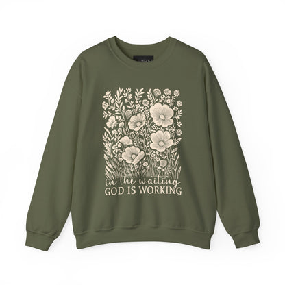 On the front of this custom women's Christian pullover sweatshirt are a bunch of wildflowers with the words in the waiting God is working underneath the flowers.
