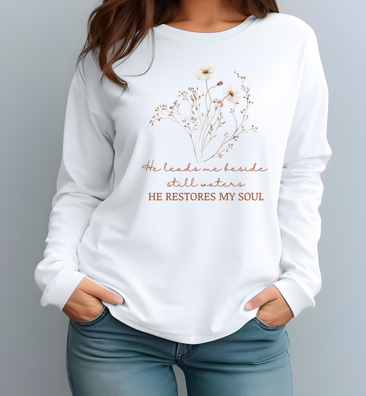 On the front of this women's Christian long sleeve shirt are a bunch of beige and brown flowers and leaves on the top of the shirt and under that are the words He leads me beside still waters in a cursive font with the words He restores my soul underneath that in a modern font.