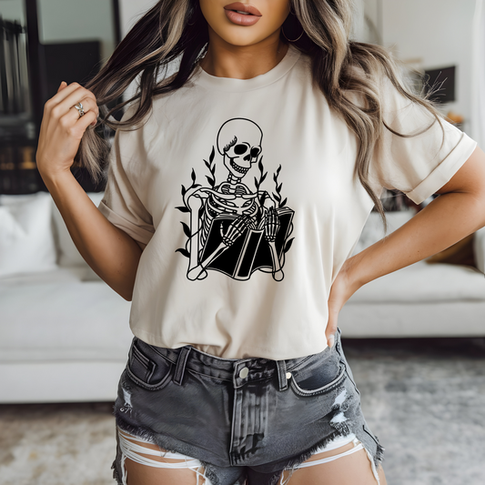 On the front of this custom unisex short sleeve shirt is a large image of a skeleton reading a book, there are vines with leaves on them behind the skeleton.
