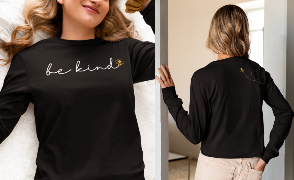 On the front of this custom unisex long sleeve shirt are the words be kind in a simple cursive font, with a bumble bee on the right side of the letter kind, on the back right shoulder there is an image of a bumble bee flying and leaving a trail behind it in the shape of a heart.