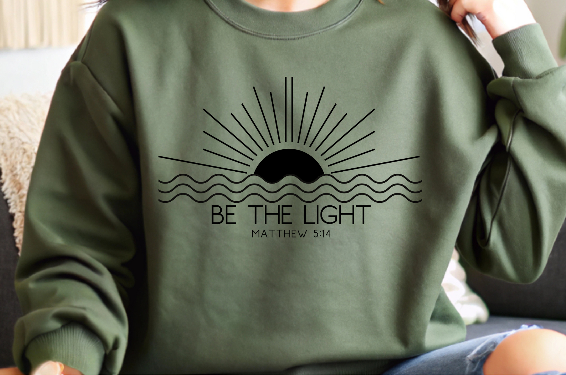 This cozy Christian crewneck sweatshirt features a sunrise over the ocean in the background with the bible verse Be the Light Matthew 5:14 in front of the image.