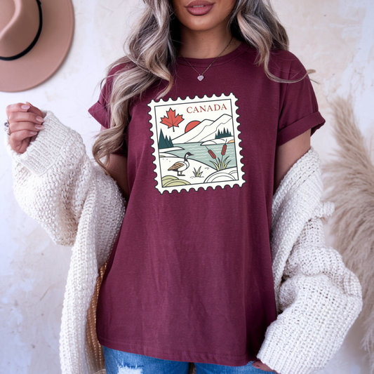 On the front of this custom short sleeve shirt is an image of a Canadian postage stamp, there is a maple leaf, the rocky mountains, a lake and a Canadian goose on the postage stamp.