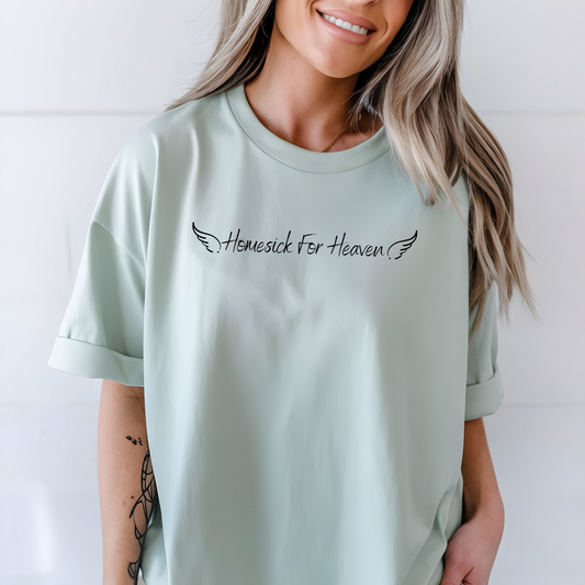 On the front of this custom vintage Christian short sleeve shirt are the words Homesick for Heaven with Angel wings on either side of the writing.