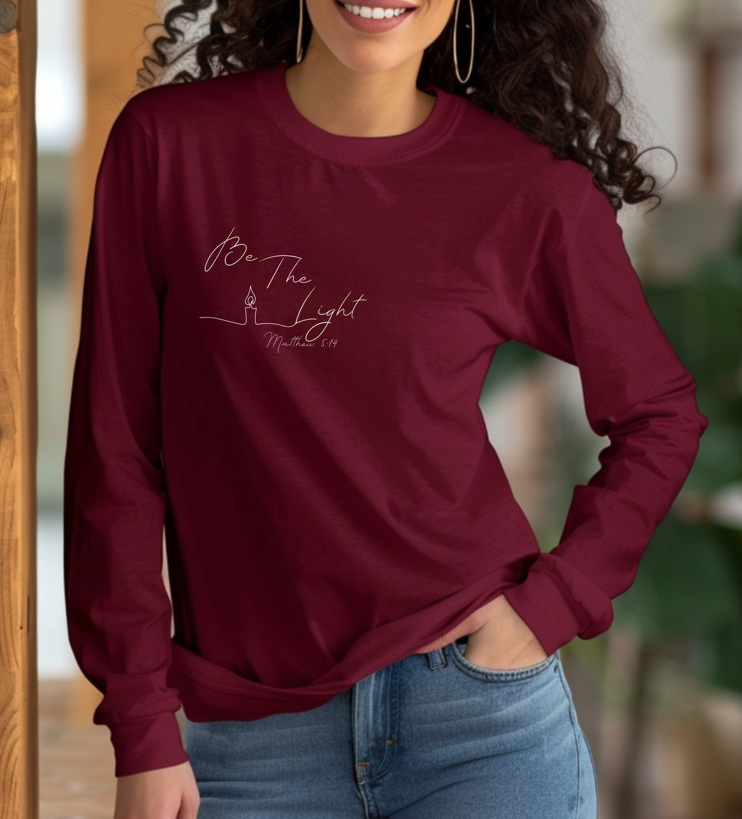 This Christian unisex Long Sleeve t-shirt features the words Be The Light Matthew 5:14 in a simple cursive font with an image of a line drawing of a candle next to the word light.