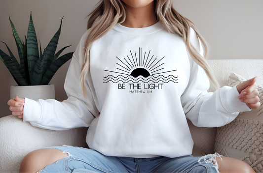 This cozy Christian crewneck sweatshirt features a sunrise over the ocean in the background with the bible verse Be the Light Matthew 5:14 in front of the image.