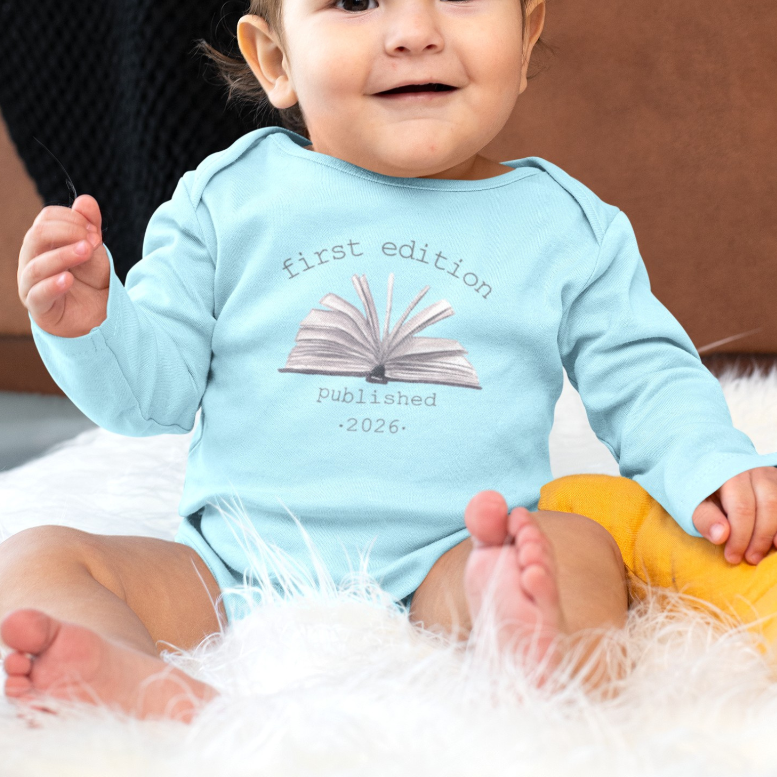 Baby wearing a light blue onesie with text and an open book graphic, sitting on a fluffy white surface.