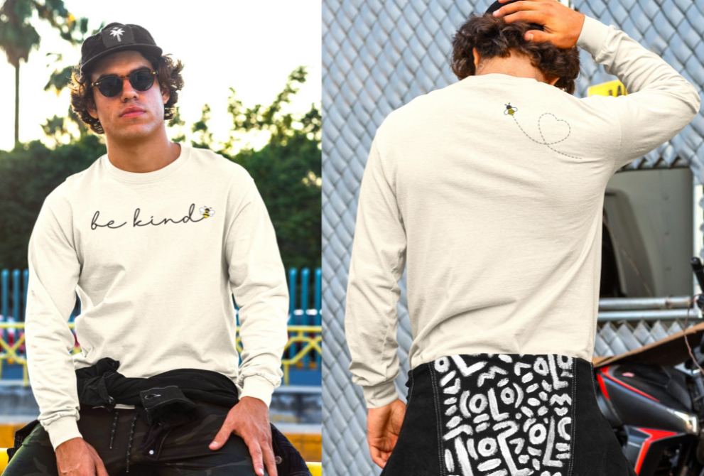 On the front of this custom unisex long sleeve shirt are the words be kind in a simple cursive font, with a bumble bee on the right side of the letter kind, on the back right shoulder there is an image of a bumble bee flying and leaving a trail behind it in the shape of a heart.