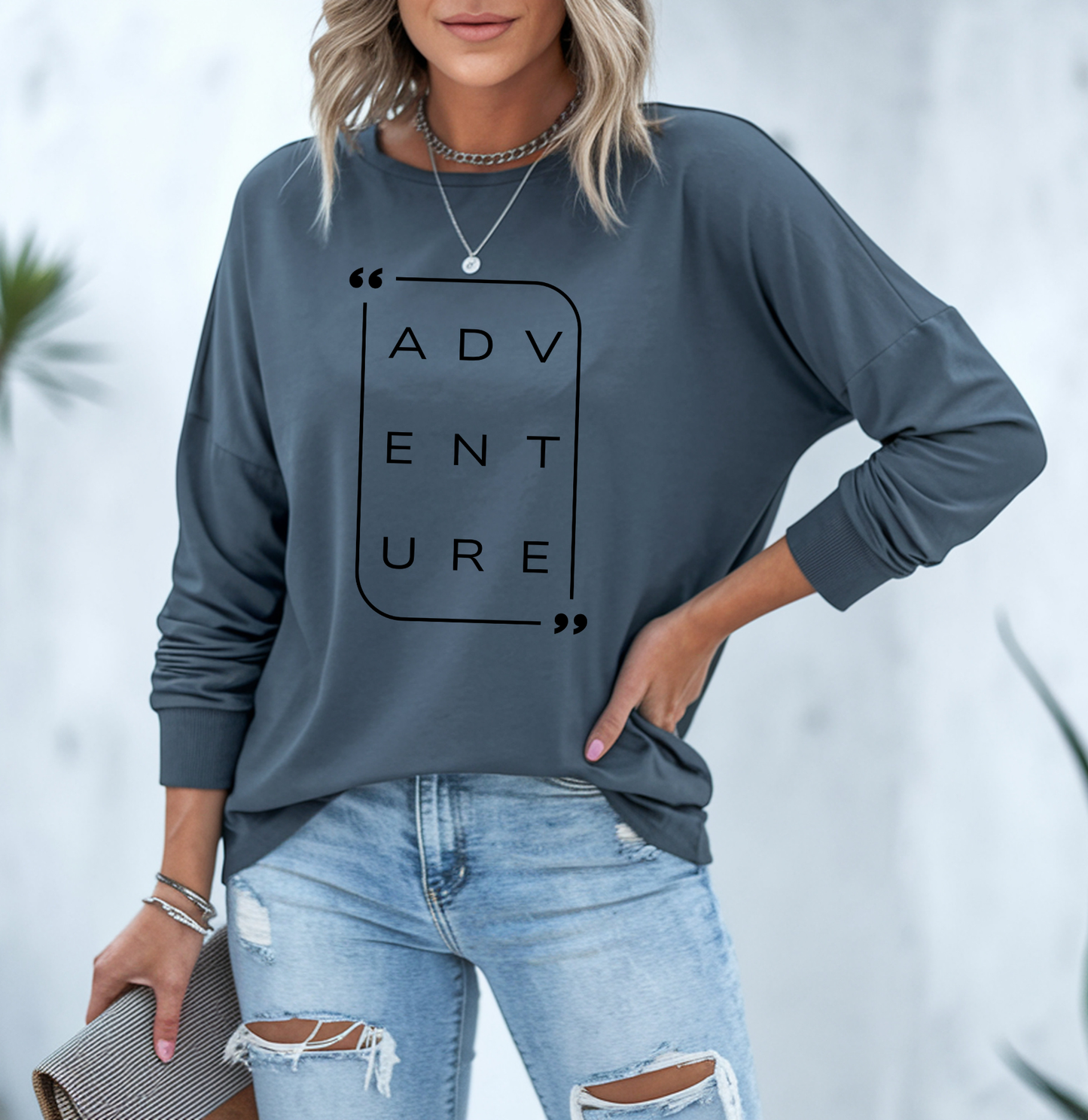 On the front of this super soft unisex long sleeve shirt is the word adventure with there being three letters on each line, it is in a modern font. There is a border around the word.