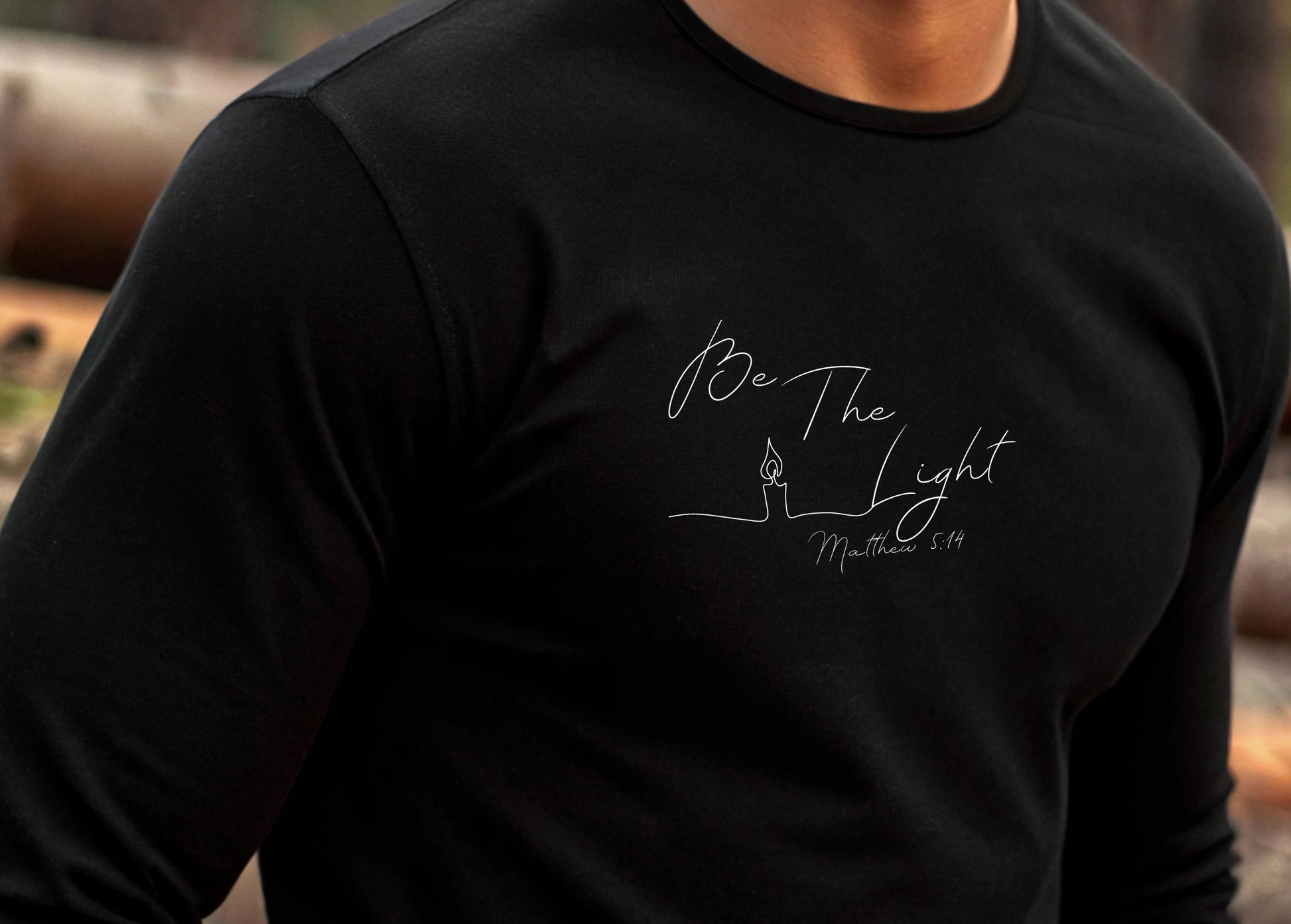 This Christian unisex Long Sleeve t-shirt features the words Be The Light Matthew 5:14 in a simple cursive font with an image of a line drawing of a candle next to the word light.