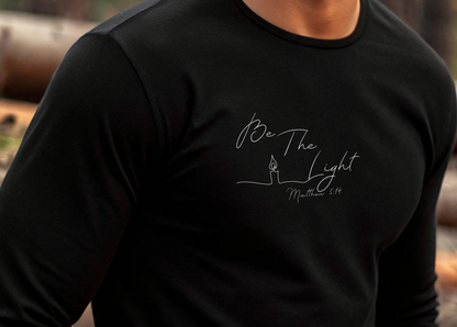 This Christian unisex Long Sleeve t-shirt features the words Be The Light Matthew 5:14 in a simple cursive font with an image of a line drawing of a candle next to the word light.