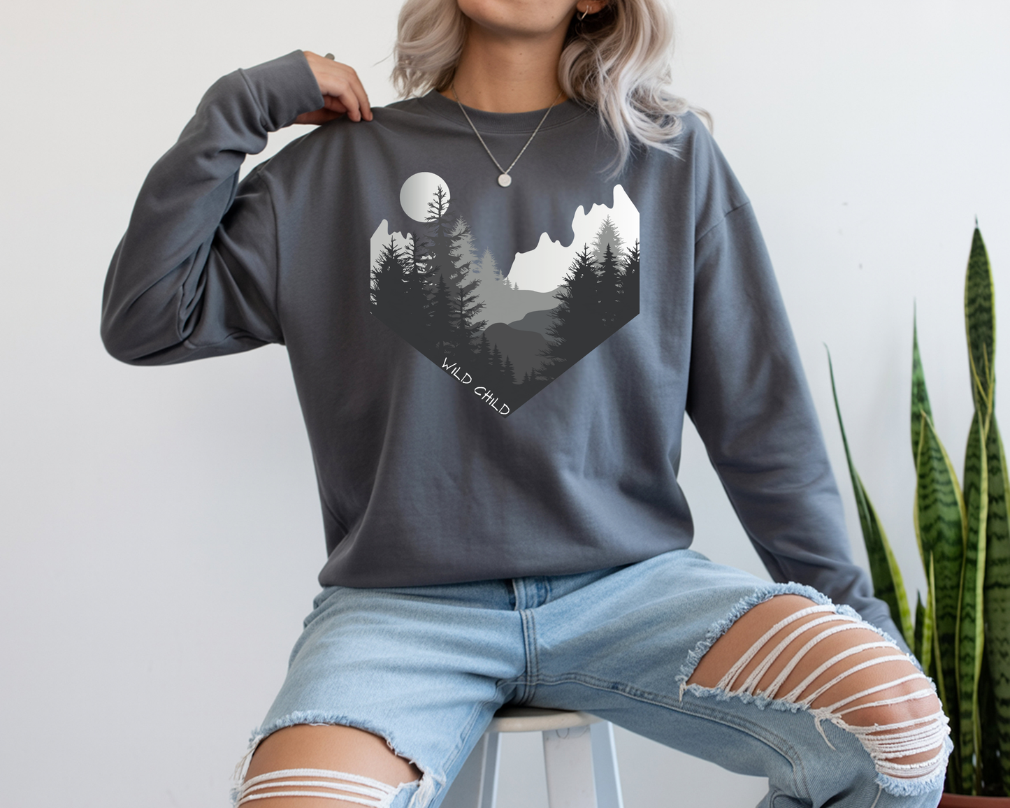 The image on the front of this custom long sleeve shirt is all in black and grey with there being mountains and trees on both sides in the shape on a v making it looking as if you are looking down a valley, the words wild child are in white on the bottom left of the image.
