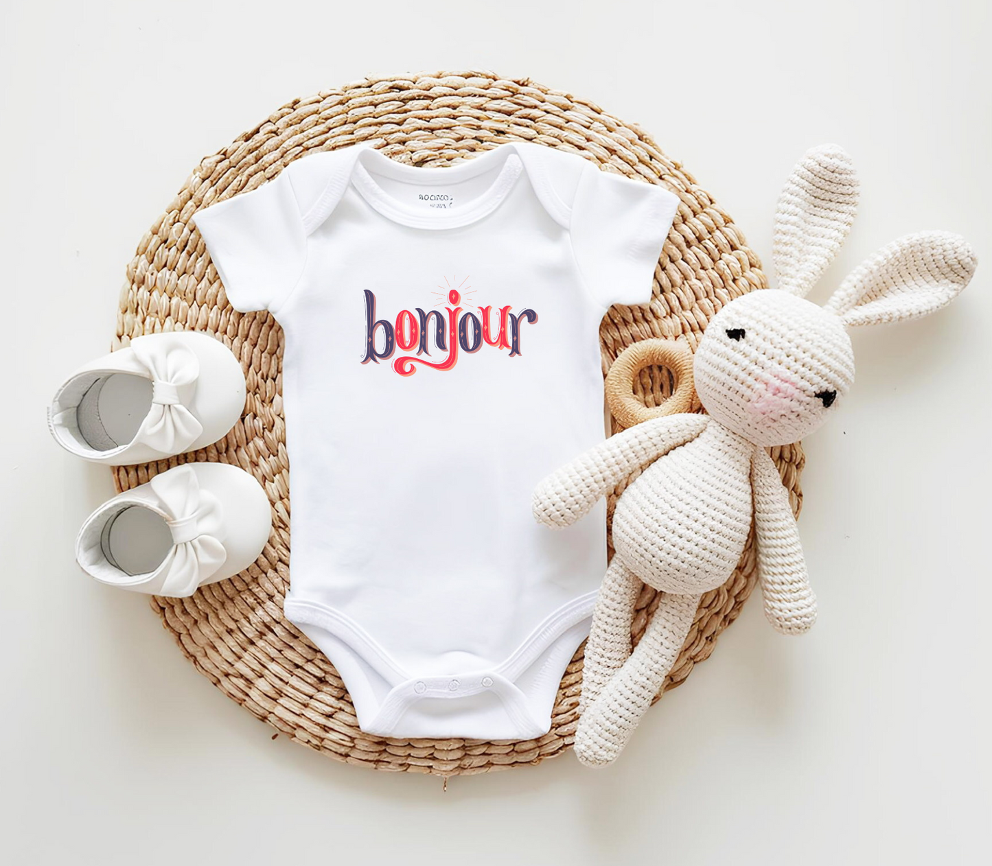 A custom infant onesie that has shirt sleeves and three buttons on the bottom, across the front of this custom baby bodysuit is the word bonjour in blue and red writing.