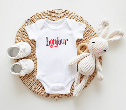 A custom infant onesie that has shirt sleeves and three buttons on the bottom, across the front of this custom baby bodysuit is the word bonjour in blue and red writing.