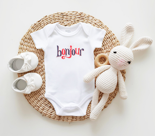 A custom infant onesie that has shirt sleeves and three buttons on the bottom, across the front of this custom baby bodysuit is the word bonjour in blue and red writing.