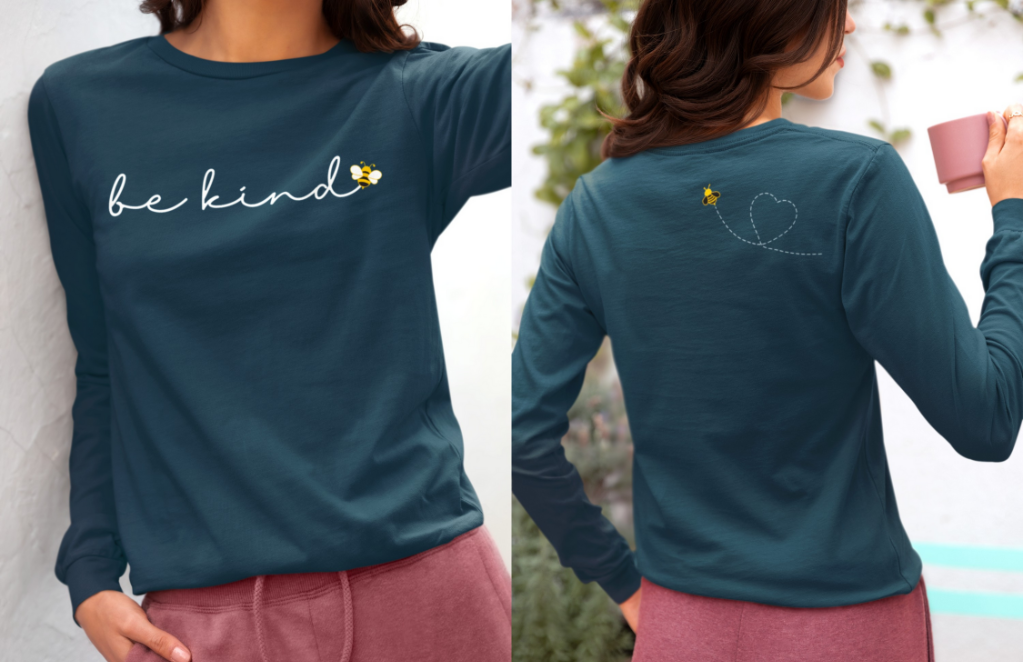 On the front of this custom unisex long sleeve shirt are the words be kind in a simple cursive font, with a bumble bee on the right side of the letter kind, on the back right shoulder there is an image of a bumble bee flying and leaving a trail behind it in the shape of a heart.