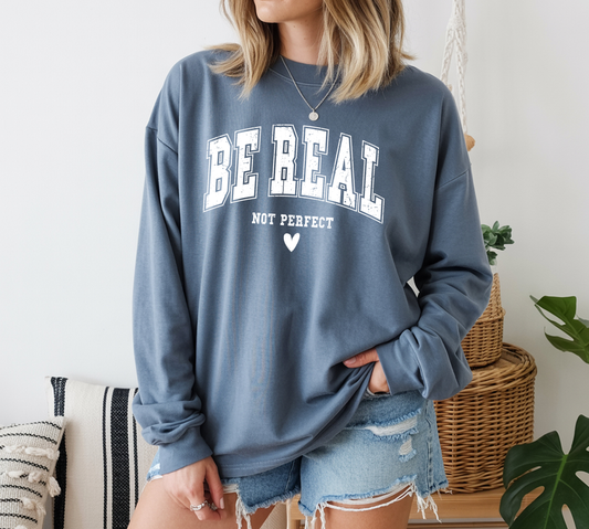 On the front of this custom unisex long sleeve shirt are the words Be Real in large bubble font with the words not perfect underneath the bubble letters, on the bottom there is a heart.