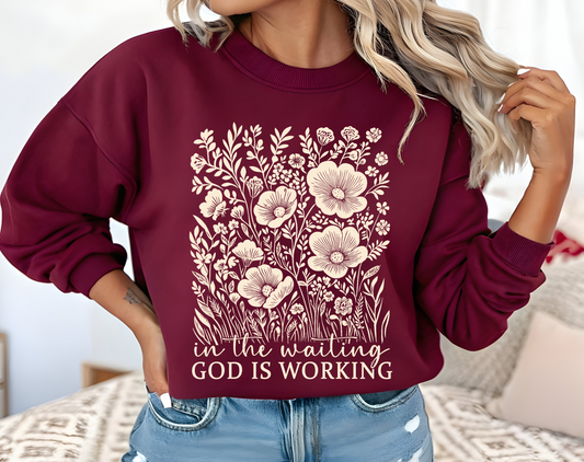 On the front of this custom women's Christian pullover sweatshirt are a bunch of wildflowers with the words in the waiting God is working underneath the flowers.