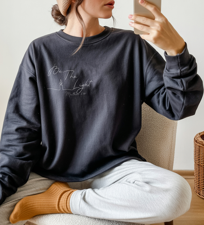 This Christian unisex Long Sleeve t-shirt features the words Be The Light Matthew 5:14 in a simple cursive font with an image of a line drawing of a candle next to the word light.