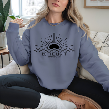 This cozy Christian crewneck sweatshirt features a sunrise over the ocean in the background with the bible verse Be the Light Matthew 5:14 in front of the image.
