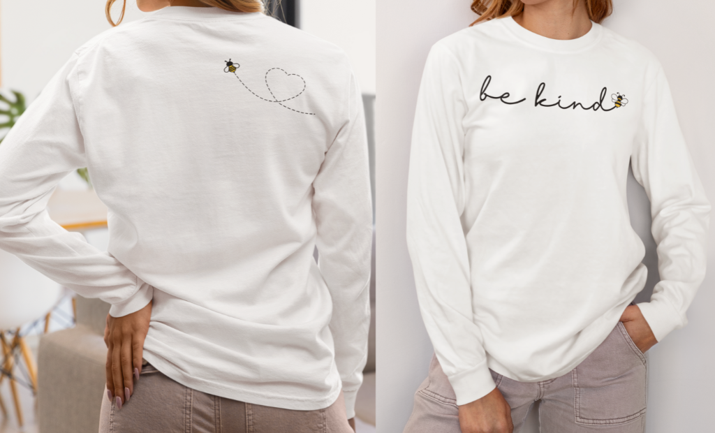 On the front of this custom unisex long sleeve shirt are the words be kind in a simple cursive font, with a bumble bee on the right side of the letter kind, on the back right shoulder there is an image of a bumble bee flying and leaving a trail behind it in the shape of a heart.