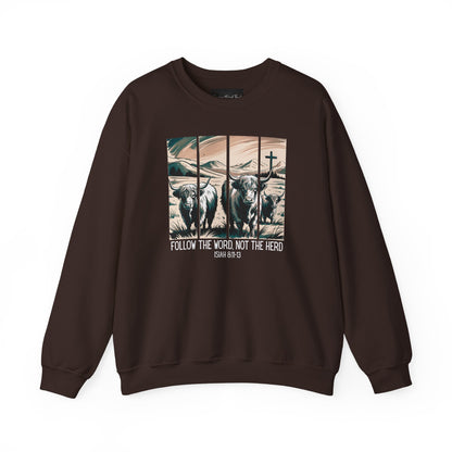 On the front of this Christian pullover sweatshirt is the image of cows standing in a pasture that is in a valley surrounded by mountains, there is a Jesus cross behind the cows, underneath the image is the scripture Follow the word, not the herd Isiah 8:11-13