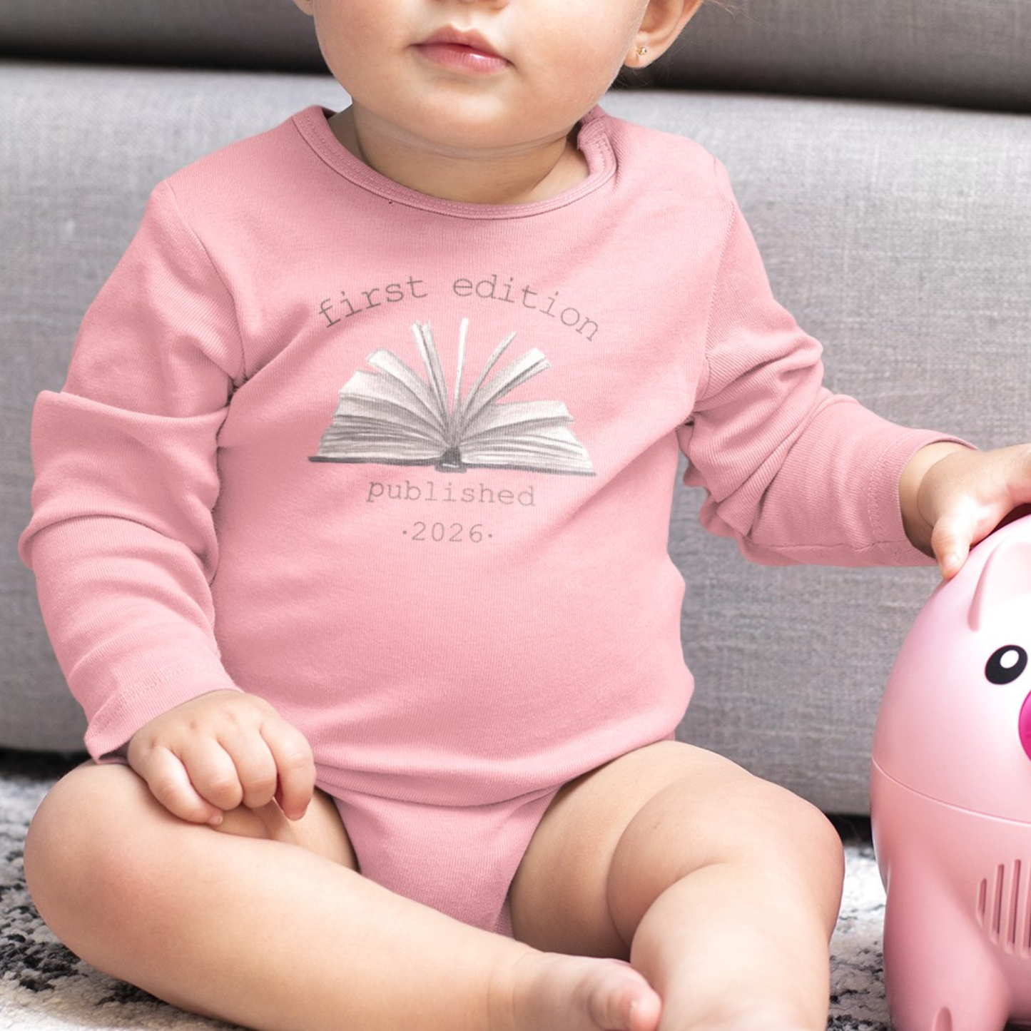 On the front of this custom baby onesie are the words First Edition Published 2026 with an image of an open book in the middle of the wording.