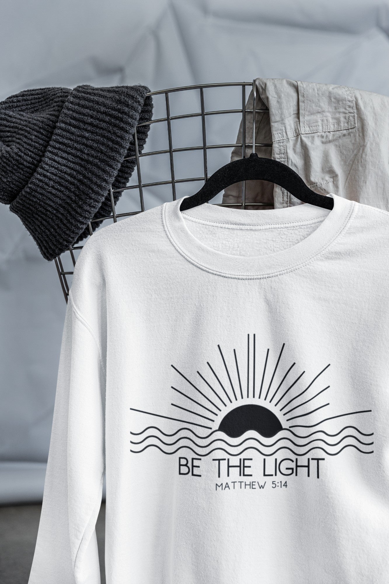 This cozy Christian crewneck sweatshirt features a sunrise over the ocean in the background with the bible verse Be the Light Matthew 5:14 in front of the image.