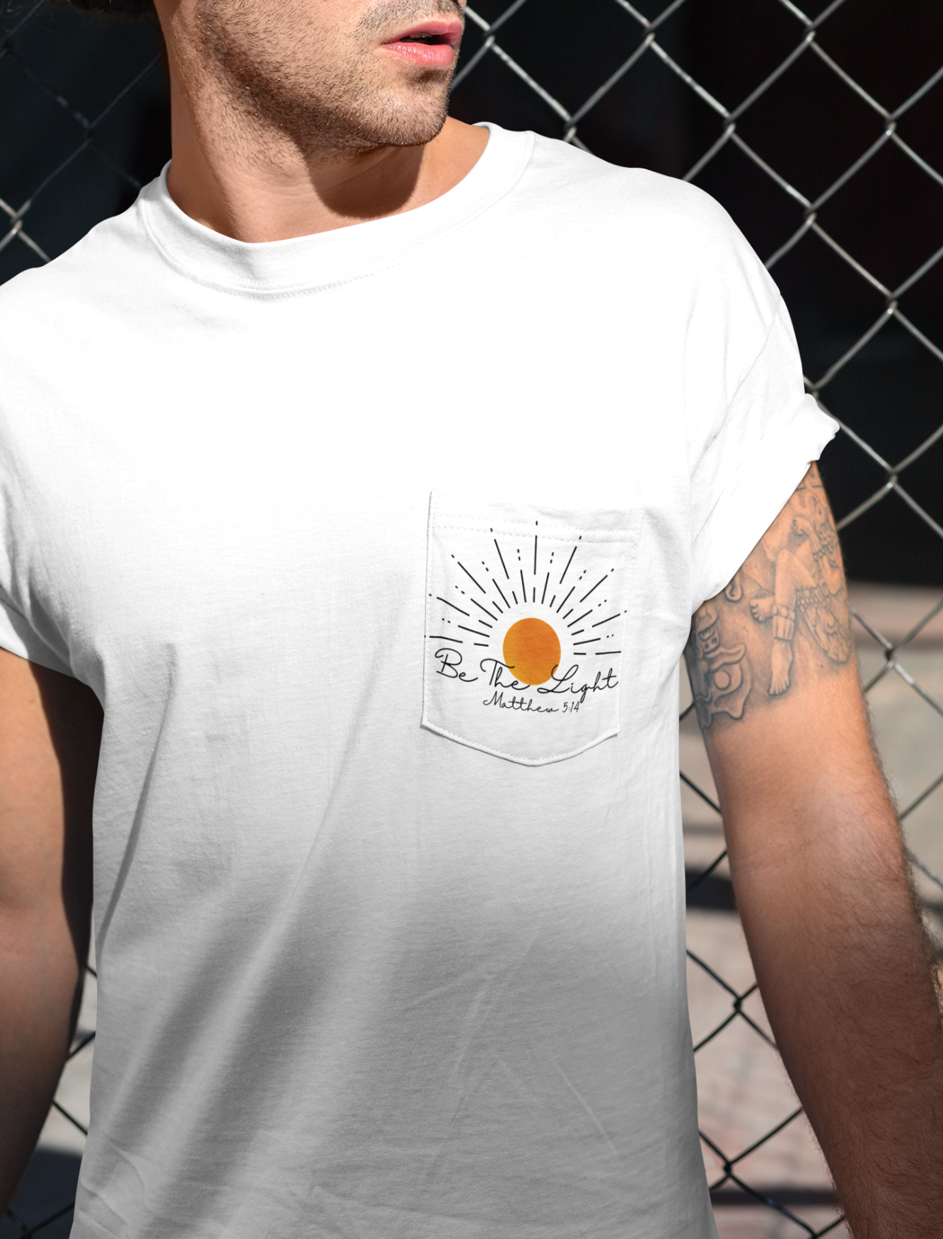 A custom Christian unisex pocket t-shirt, on the pocket that is over the left chest of the shirt is a large sun with the words Be The Light Matthew 5:14 of the Bible.