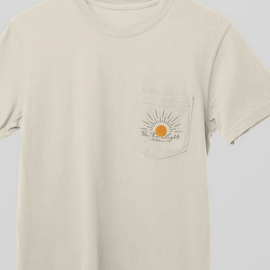 A custom Christian unisex pocket t-shirt, on the pocket that is over the left chest of the shirt is a large sun with the words Be The Light Matthew 5:14 of the Bible.